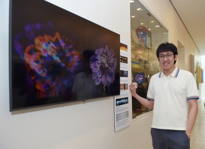 Ethan Tseng standing in the hallway of the Friend Center next to a large TV monitor displaying 2 images of flowers side by side.  The flower on the right is a clearer image.  Ethan is holding a small postcard of the clearer flower.