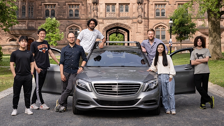 Seven people posing with a car outdoors. The car is gray and the doors are open.