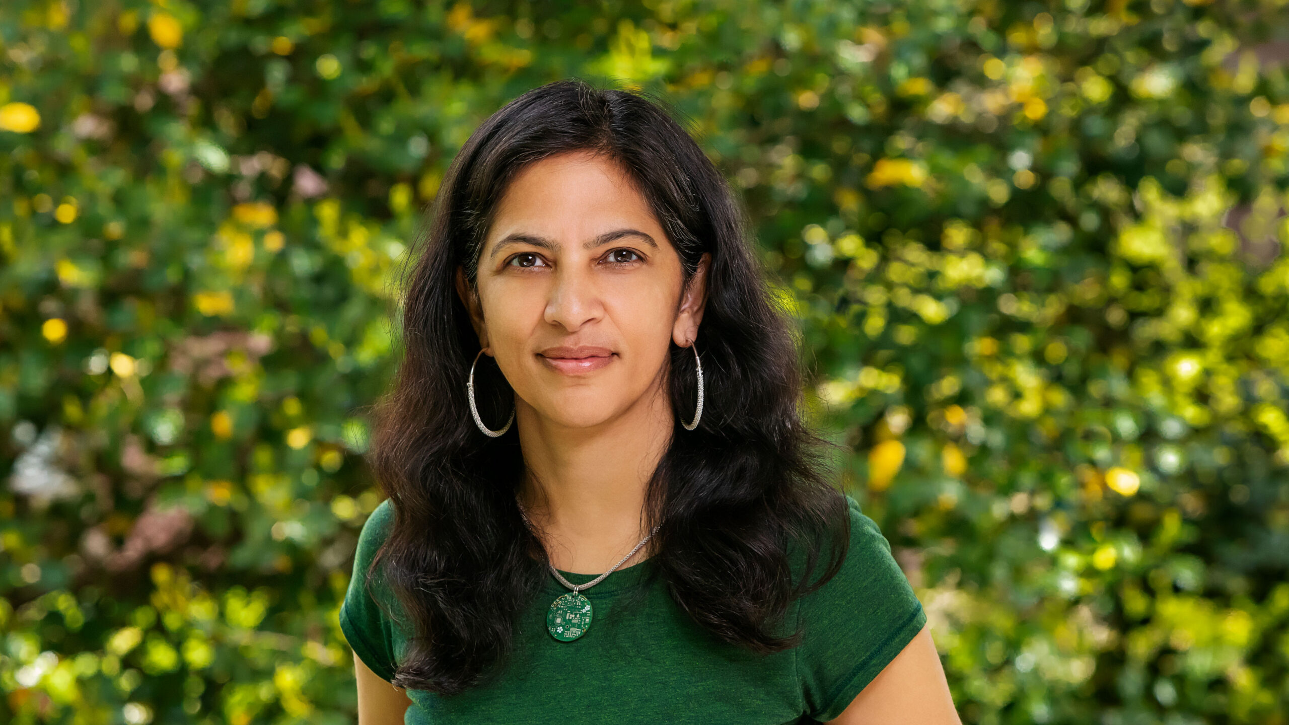 Head and shoulders photo of Radhika Nagpal in a green t-shirt standing in front of green foliage.