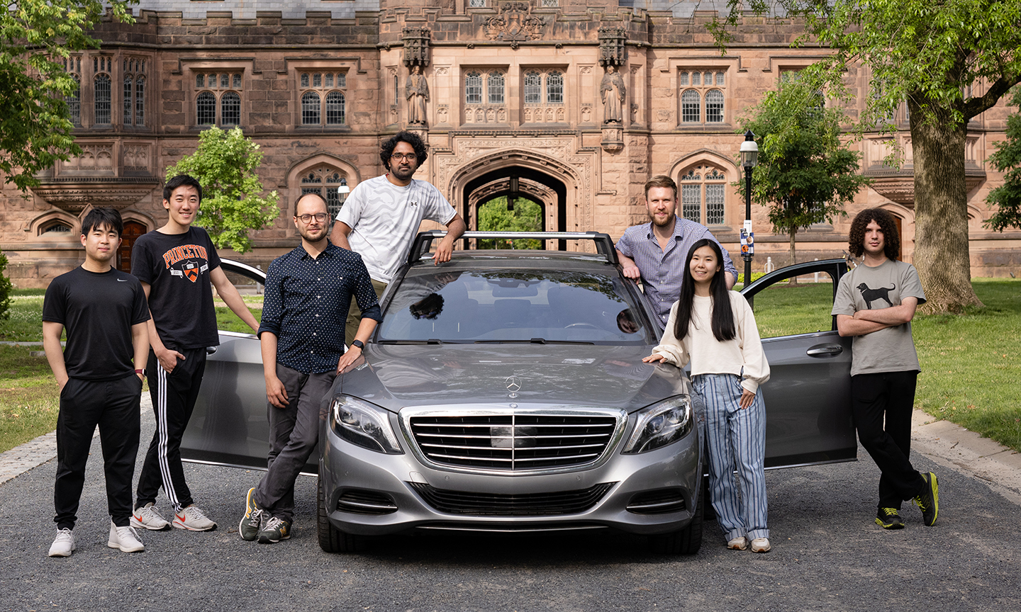 Heide's lab group standing outside on Princeton's campus posing with a test vehicle.  The car's doors are open and it's parked on a gravel road.