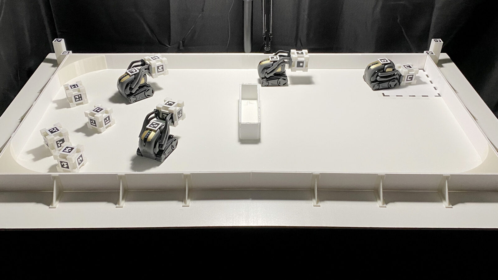 Four tiny robots on a table top with smaller marked cubes.  The robots are moving the cubes from the left side to a marked spot on the right.