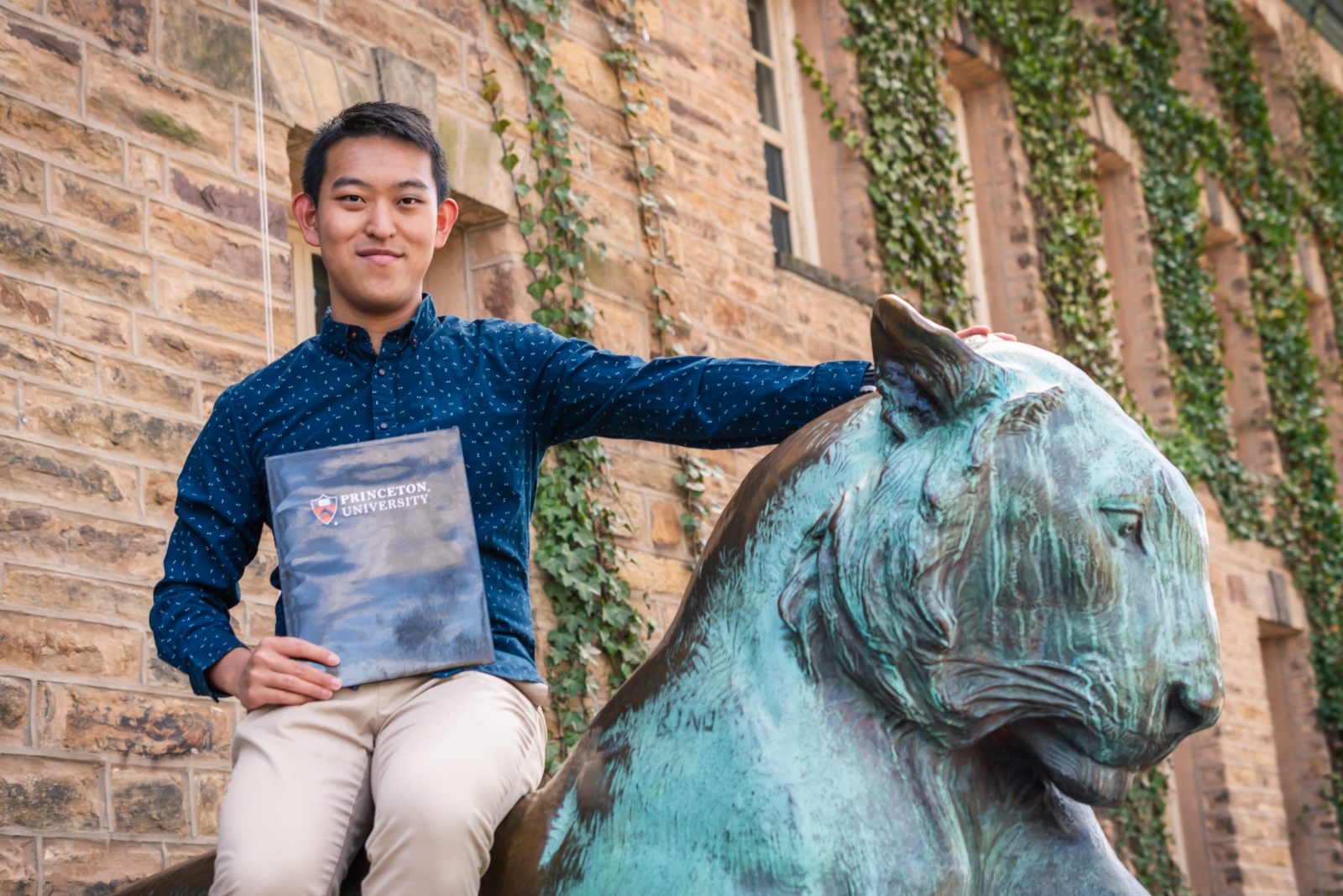 Yang Song sitting on the bronze tiger on the front steps of Nassau Hall.  He's holding a black folder with the Princeton University logo on the front.